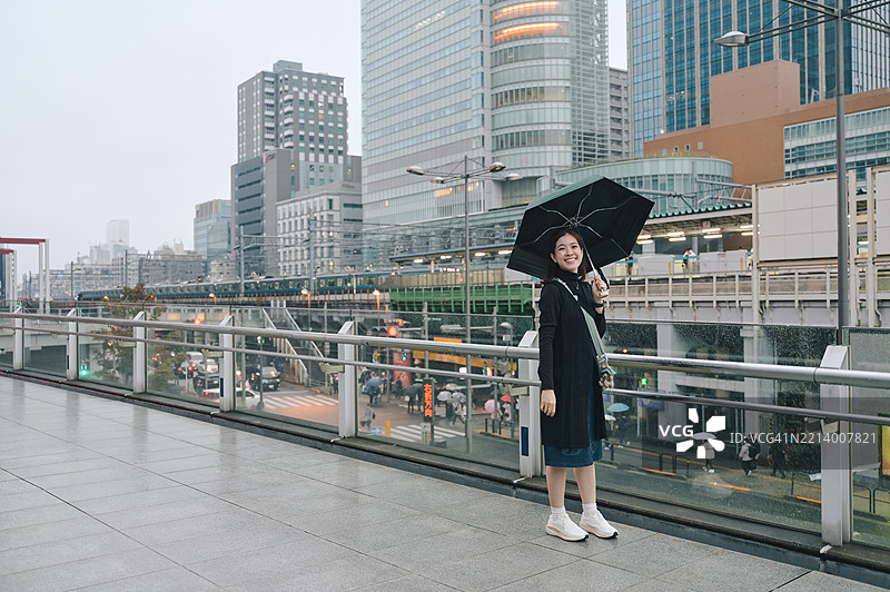 年轻的日本女性微笑着手持雨伞，背景是黄昏时分繁忙的东京新宿市区。图片素材