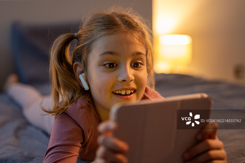 微笑的年轻女孩在温馨的卧室里使用数字平板，柔和的晚间灯光洒满房间。图片素材