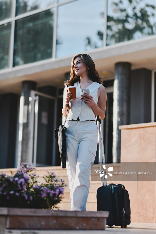 女性手持咖啡杯、行李箱和手机，等待朋友时站着图片素材