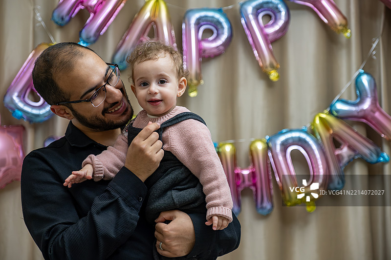 父亲抱着宝宝庆祝生日，背景中有五彩缤纷的气球图片素材