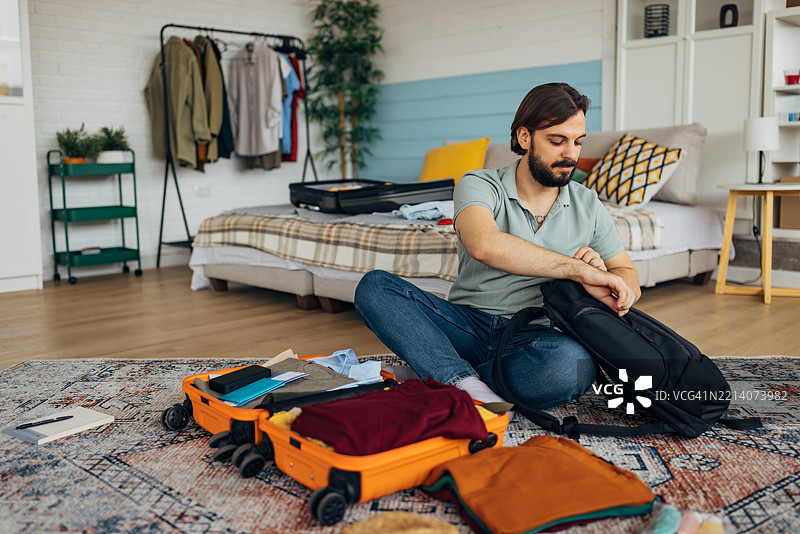 一个无忧无虑的男人正在为旅行打包。他坐在卧室的地板上，整理一个行李箱和一个背包。图片素材