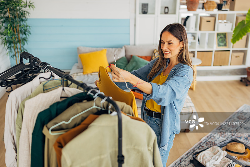 一位美丽微笑的女性站在衣架旁，正在挑选旅行要带的衣物。图片素材