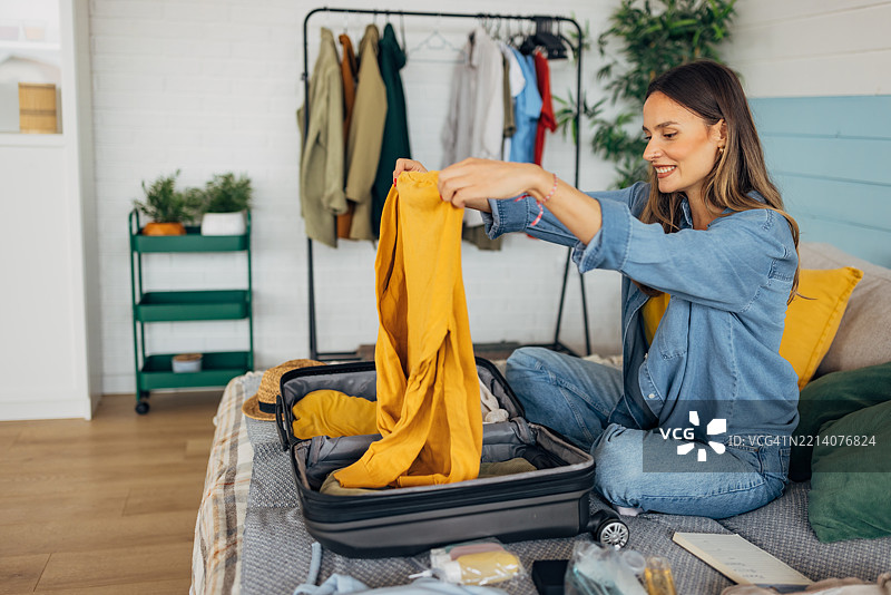 女人在家里的床上把衣服装进行李箱，微笑着享受整理行李的过程，为一次激动人心的旅行做准备。图片素材