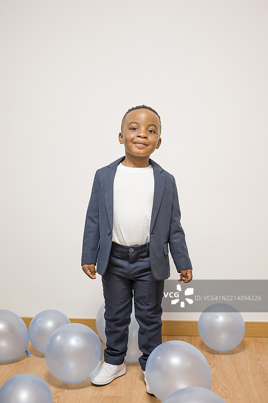 微笑的年轻男孩穿着时尚的海军蓝西装和白衬衫，自豪地站在气球中间。图片素材