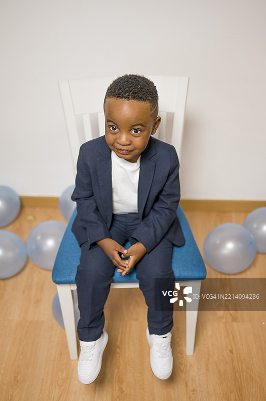 小男孩安静地坐在白色椅子上，穿着深蓝色西装和运动鞋。图片素材