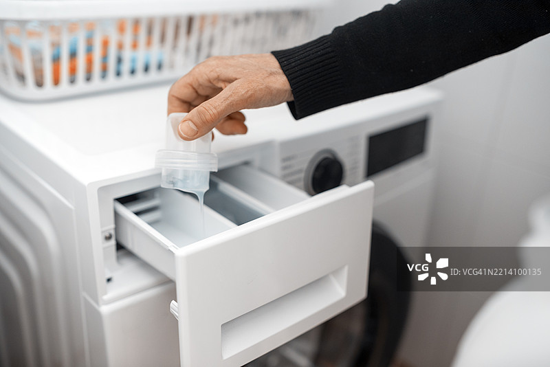 家庭中一位女性的手特写，正在将柔顺剂或洗衣液倒入洗衣机中清洗衣物。图片素材