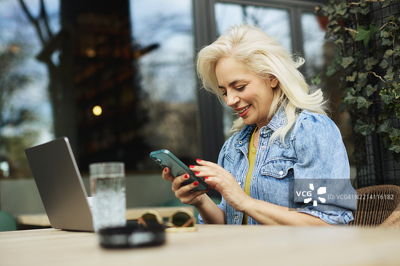 一位五十多岁的美丽女商人在咖啡馆的花园里坐着，一边喝咖啡，一边用手机写信息，同时在笔记本电脑上工作。图片素材