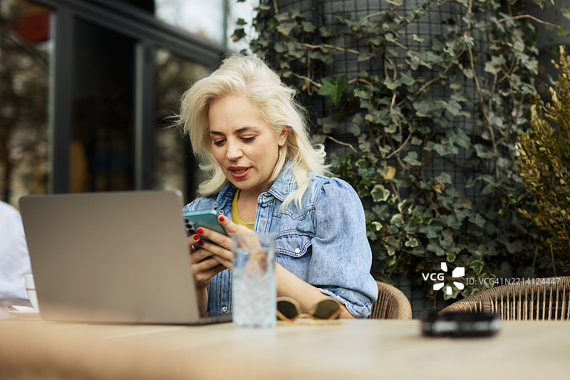 一位五十多岁的美丽女商人在咖啡馆的花园里坐着，一边喝咖啡，一边用手机写信息，同时在使用笔记本电脑。图片素材