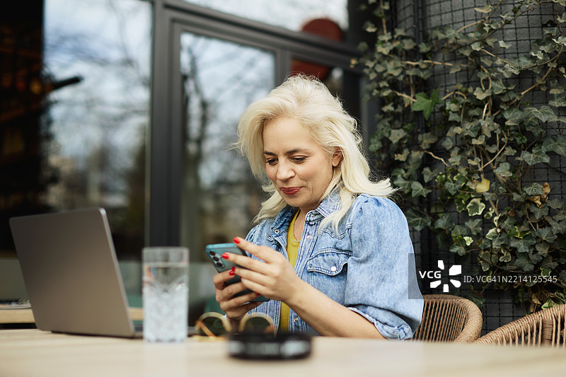 一位五十多岁的美丽女商人在咖啡馆的花园里坐着，一边喝咖啡，一边用手机写信息，同时在使用笔记本电脑。图片素材