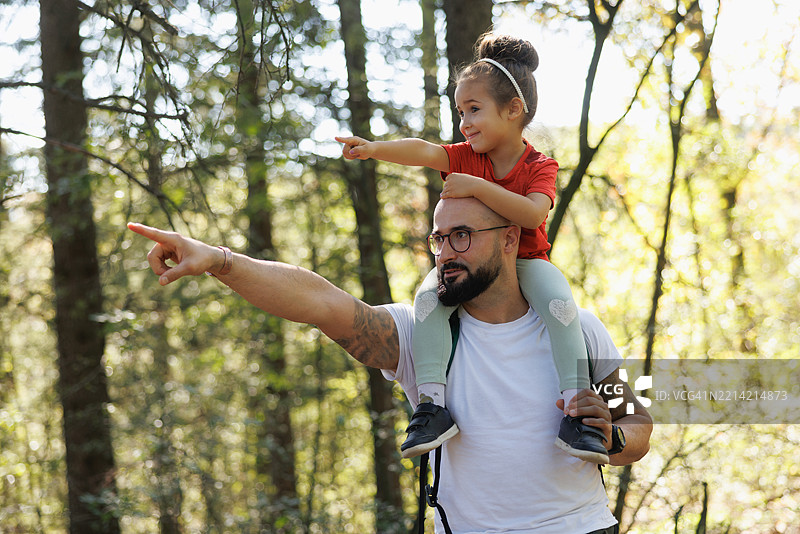 男人在美丽的日子里把小女儿扛在肩上，在树林里指着天空。图片素材