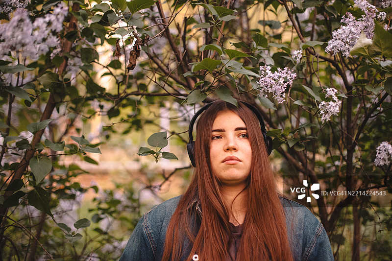 年轻女性在户外站立，戴着耳机听音乐，周围是紫丁香花。图片素材