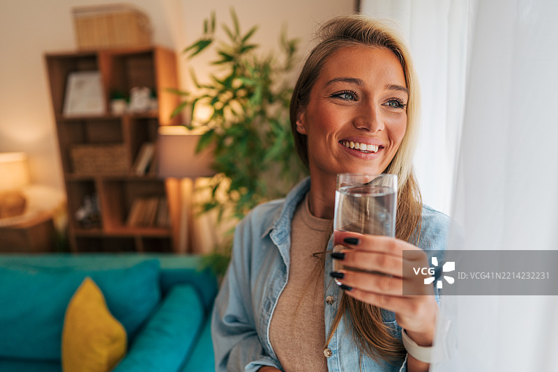 微笑的年轻女性在家中窗边喝水图片素材