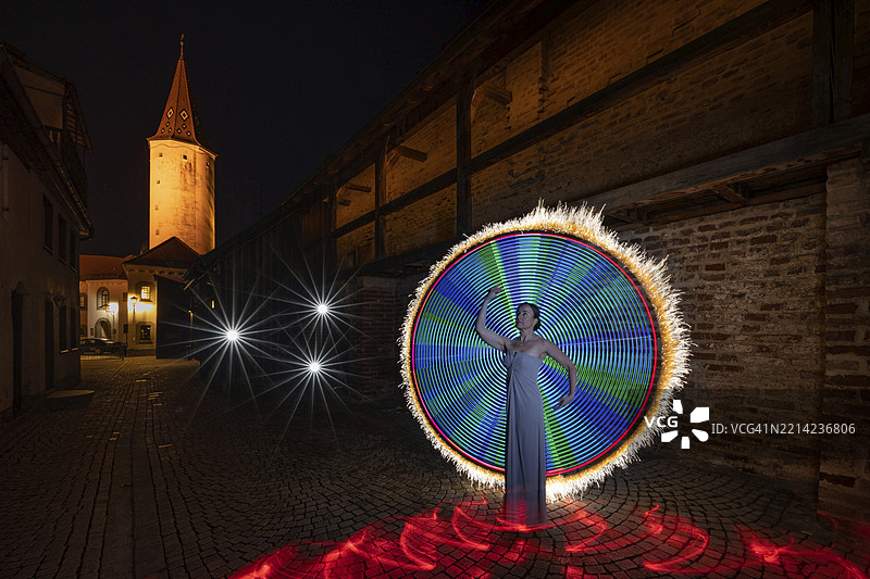 在历史悠久的老城区背景下，夜晚一条鹅卵石小巷中，一位女性站在令人印象深刻的光绘作品前，地点：德国巴伐利亚州的明德尔海姆，乌特阿尔高区。图片素材