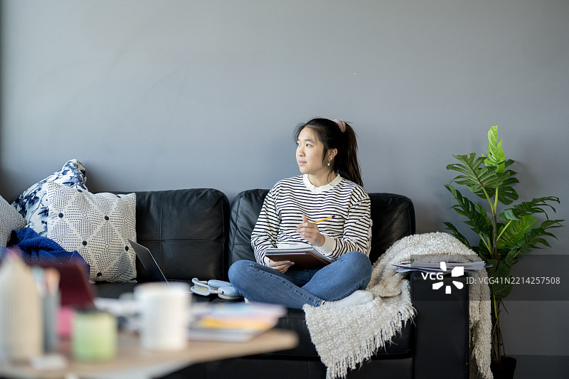 年轻女性坐在沙发上在家中写笔记本。图片素材