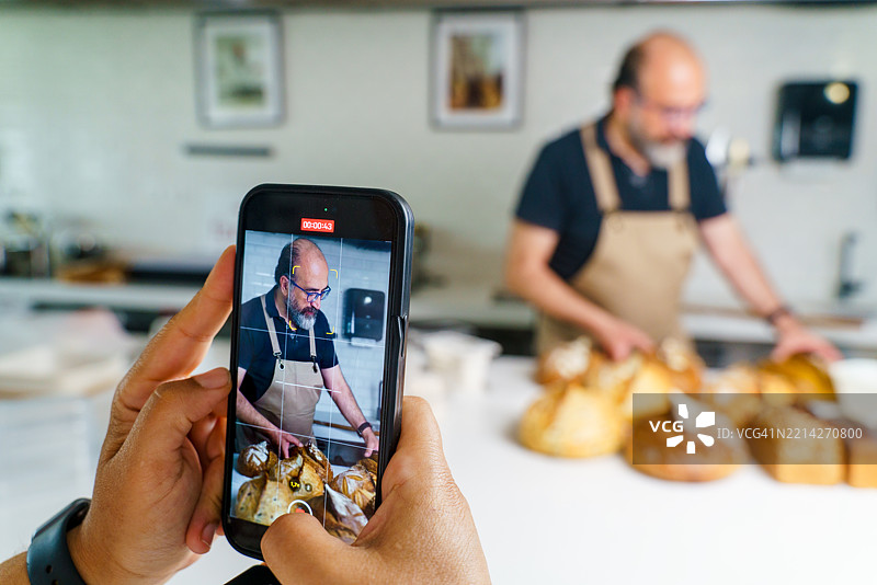面包师在手工面包店用智能手机拍摄烹饪教程图片素材