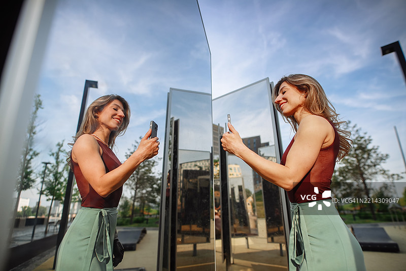 夏季公园日落：一位穿裙子的瘦女人在镜子旁欢快地跳舞，用智能手机拍摄自拍，创造难忘的瞬间。图片素材