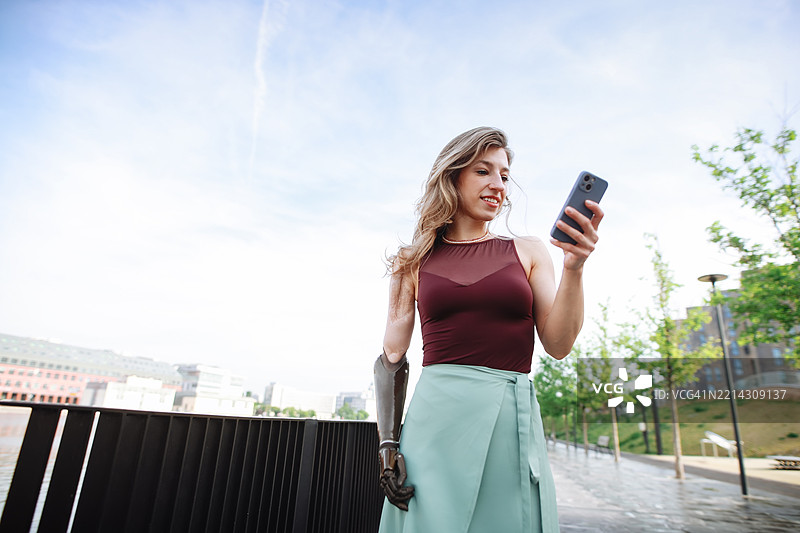 在一个宁静的城市公园里，一位美丽的女性正在与她的智能手机互动，手臂上有一个假肢。图片素材