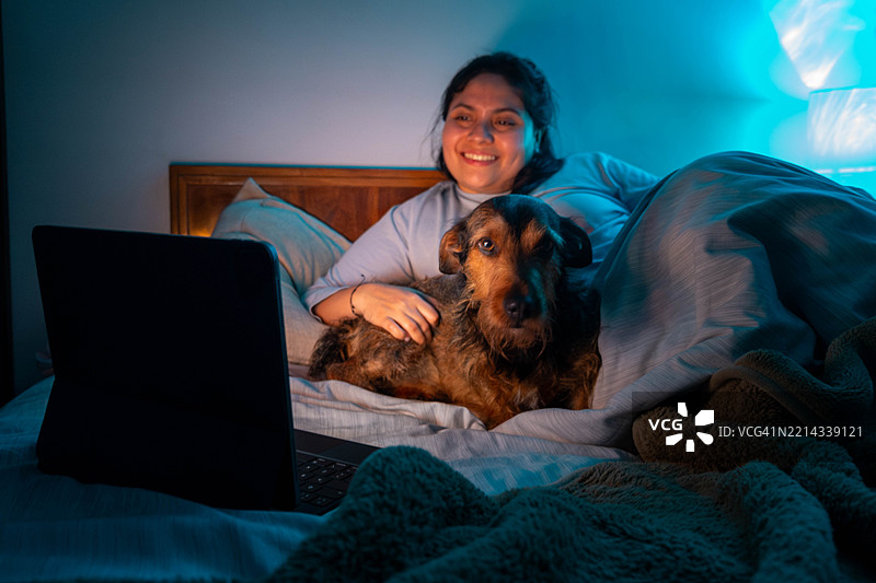 微笑的女人在床上和她的狗一起看电影，周围是温馨的氛围灯光。图片素材