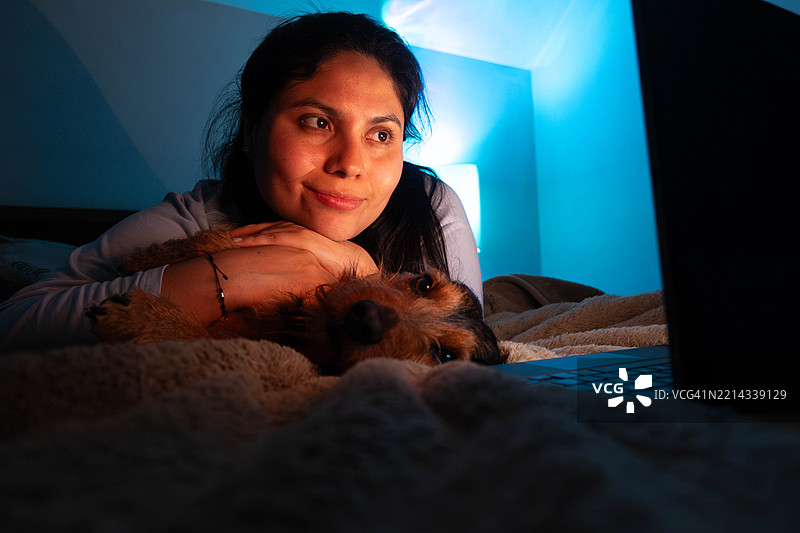舒适的电影之夜，一位女性在床上放松，与她的狗一起在柔和的蓝色灯光下观看笔记本电脑上的电影。图片素材