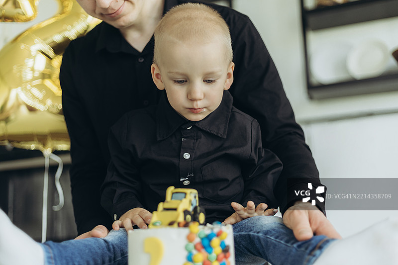 年轻男子站在一个脏兮兮的小男孩身后，小男孩坐在生日蛋糕旁。图片素材