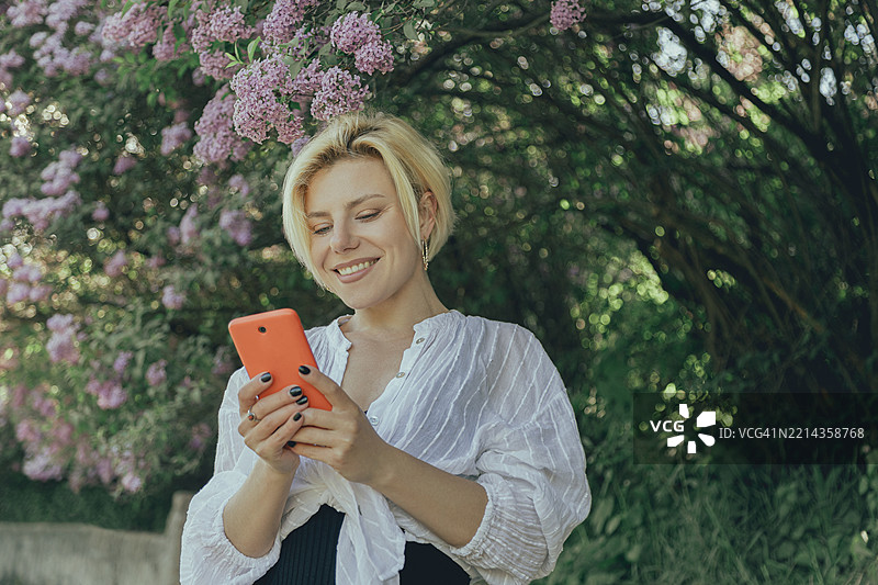 微笑的年轻女性在春天的日子里，使用智能手机，周围是盛开的紫丁香花。图片素材