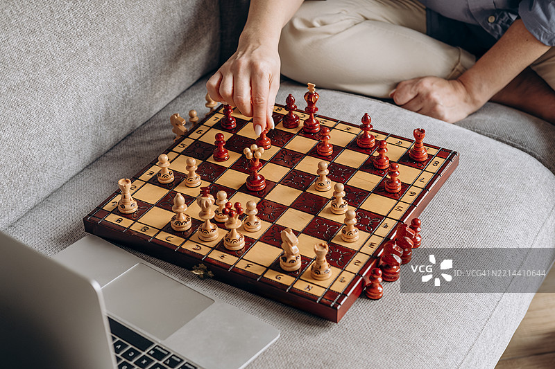 正在进行的棋局，木质棋盘上摆放着红色和米色棋子，玩家坐在舒适的沙发上进行战略性移动，旁边有一台笔记本电脑，展示出轻松的游戏环境。图片素材