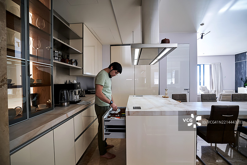 一名男子从宽敞公寓厨房岛的抽屉中拿出厨房用具。图片素材