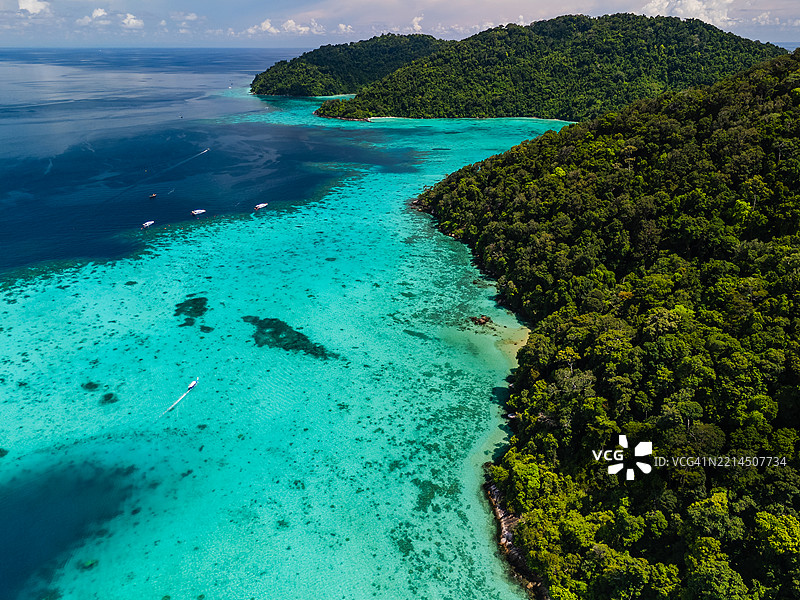 热带岛屿附近漂浮着船只的鸟瞰图，碧蓝的海水、可见的珊瑚礁和茂密的绿色丛林海岸线。图片素材