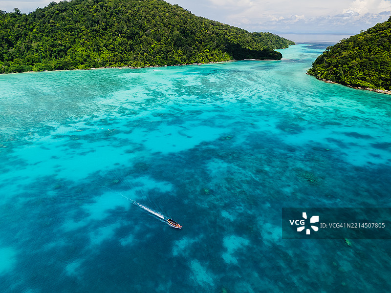 从空中俯瞰，一艘长尾船在郁郁葱葱的热带岛屿之间的鲜艳绿松石色水道中航行，下面是珊瑚礁。图片素材