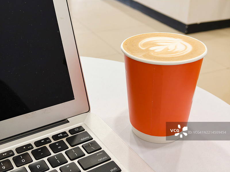 在工作桌上的橙色纸质一次性咖啡杯中盛放的拿铁艺术卡布奇诺图片素材