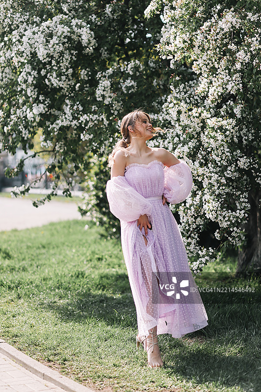 一位美丽迷人的年轻女性穿着轻盈的粉色长裙，妆容鲜艳，漫步在春夏季节的公园草坪上，享受着盛开的花朵和树木。图片素材