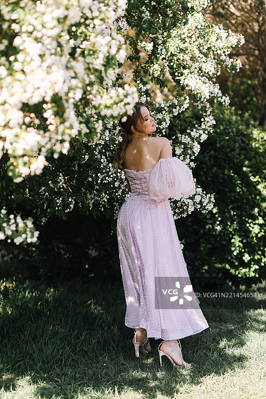 一位美丽迷人的年轻女性穿着轻盈的粉色长裙，妆容鲜艳，漫步在春夏季节的花园草坪上，享受着盛开的花朵和树木。图片素材