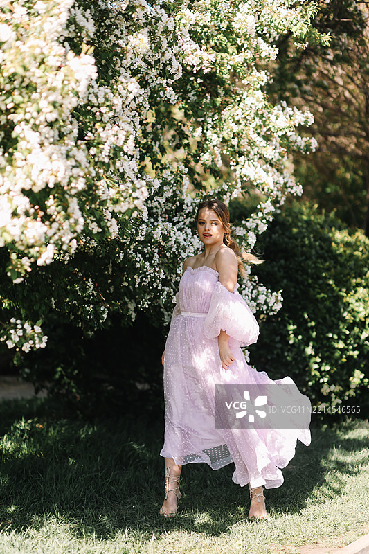 一位美丽迷人的年轻女性穿着轻盈的粉色长裙，妆容鲜艳，漫步在春夏季节的花园草坪上，享受着盛开的花朵和树木。图片素材