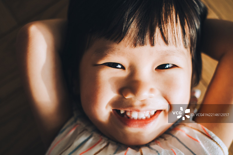 可爱的亚洲小女孩躺在家里的木地板上，双手放在头后，微笑着仰望镜头。家庭生活方式。纯真的无邪与快乐。图片素材