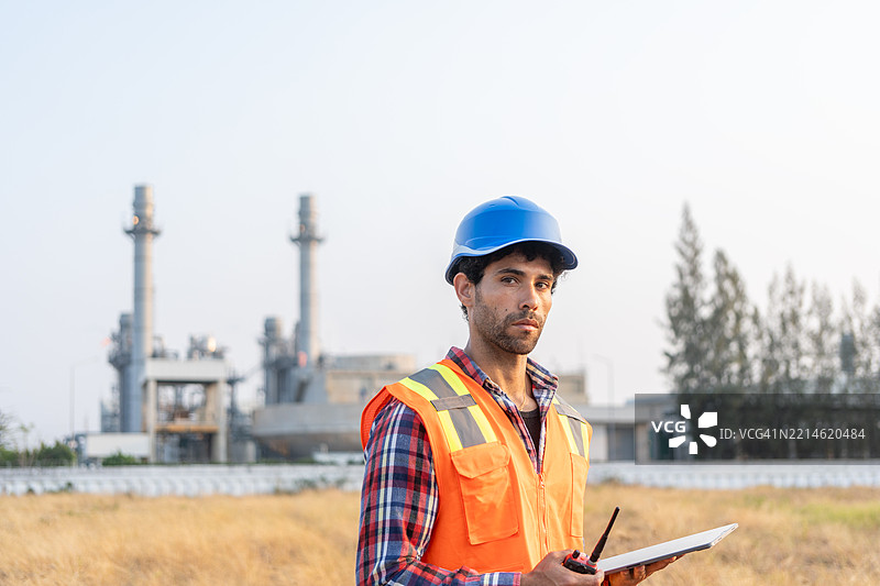 戴着安全头盔的工程师站在石油炼油厂前。工业区的天然气石化厂，油罐和管道。图片素材