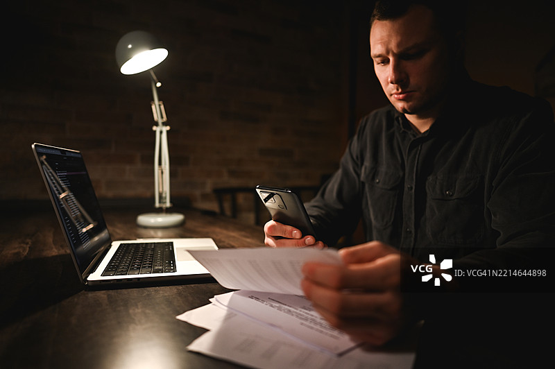 专注的商人在黑暗的家庭办公室里使用笔记本电脑和智能手机加班。图片素材