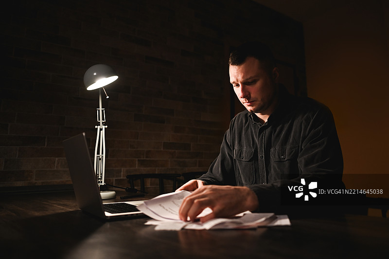 专注的商人在黑暗的办公室里加班，灯下审阅文件。图片素材