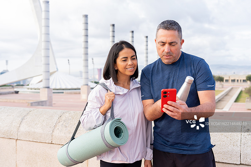 西班牙裔情侣共享智能手机屏幕，瑜伽课后面带微笑，女性手持瑜伽垫，展现锻炼后的友好氛围图片素材