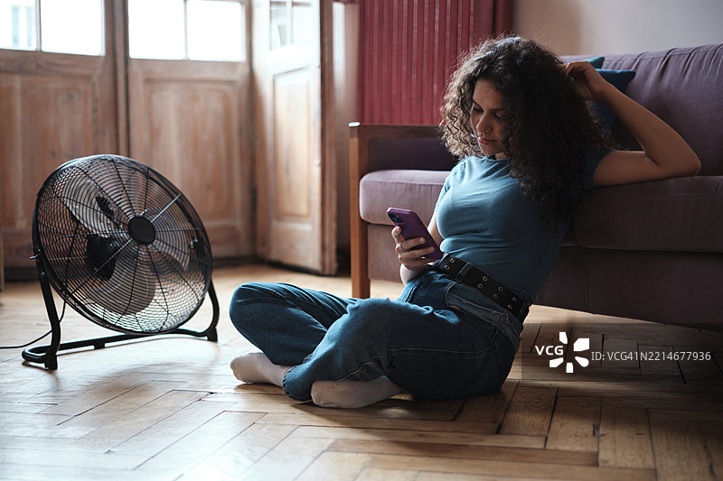 年轻女性坐在地板上，靠近电风扇，使用手机，居家生活在没有空调的公寓里。夏季炎热的概念图片素材