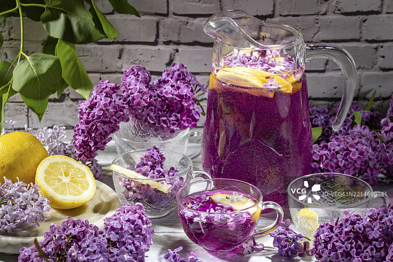 紫色鸡尾酒、柠檬片和紫丁香（Syringa）装在壶和玻璃杯中，背景是砖墙。图片素材