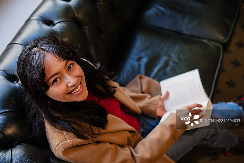年轻女性微笑着坐在切斯特菲尔德沙发上阅读一本书图片素材
