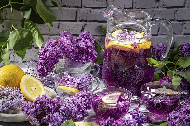 紫色鸡尾酒、柠檬片和丁香（Syringa）装饰的水壶和玻璃杯，背景是砖墙。图片素材