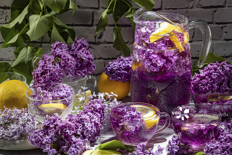 紫色鸡尾酒、柠檬片和丁香（Syringa）装饰的水壶和玻璃杯，背景是砖墙。图片素材