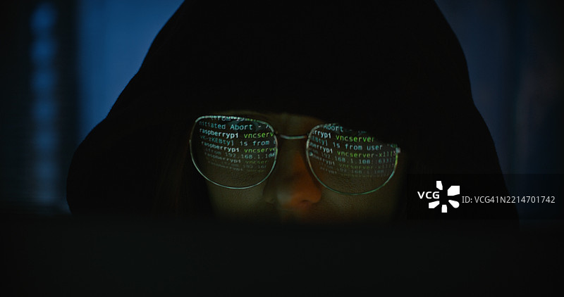 女性黑客在电脑上工作以实施网络犯罪。电脑屏幕上显示的代码反射在眼镜上。计算机犯罪图片素材