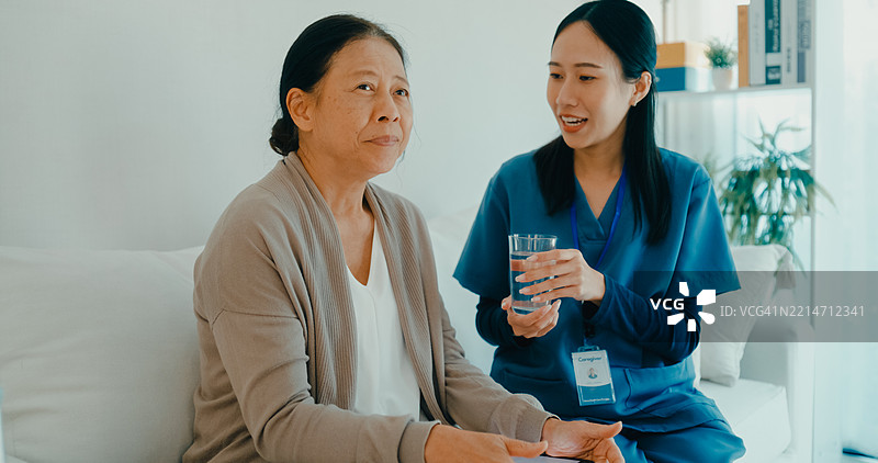 年轻的亚洲护士在家中的沙发上帮助一位老年女性服药，提供关怀和支持。家庭健康护理。图片素材