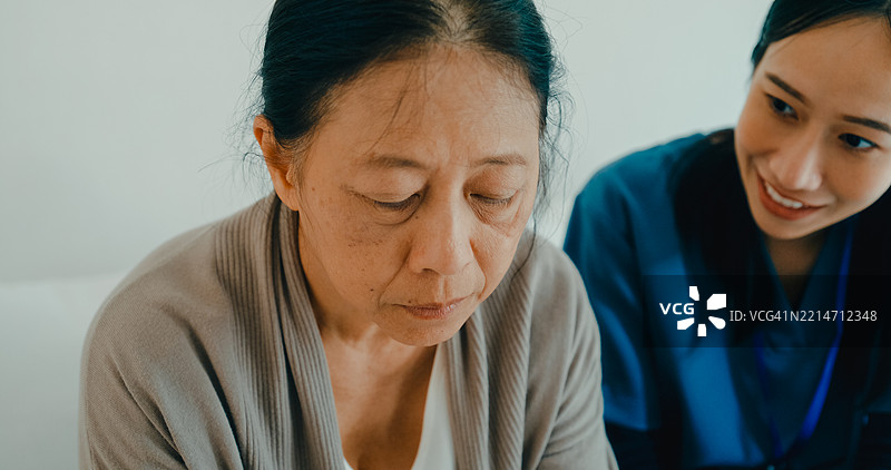年轻的亚洲护士在沙发上安慰一位老年女性，提供情感支持和关怀，家庭健康护理。图片素材