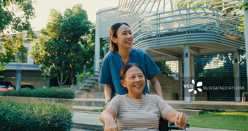 年轻的亚洲护士在公园里推着微笑的老年女性轮椅，传递支持与快乐。户外健康护理概念。图片素材