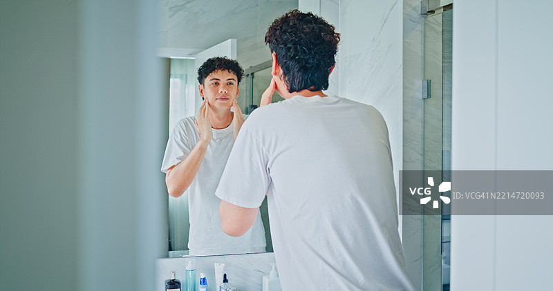 年轻男子在家浴室的镜子前微笑着涂抹面霜。健康生活方式的早晨日常概念。图片素材