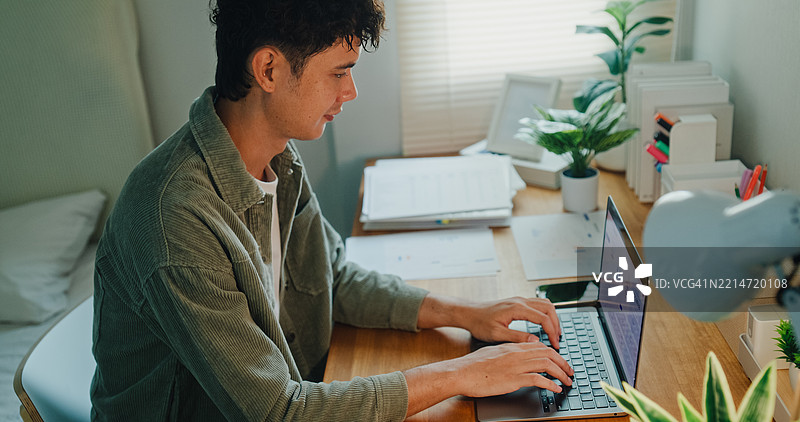 年轻的亚洲男性在家中工作，正在卧室里用笔记本电脑分析财务数据，体现现代家庭和远程工作的场景。远程工作与金融。图片素材