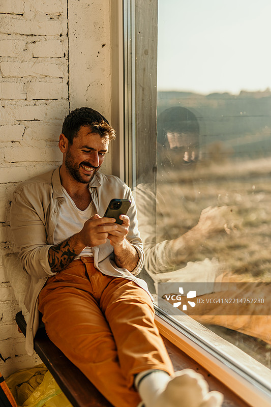 年轻男子在窗边放松，使用智能手机并微笑图片素材